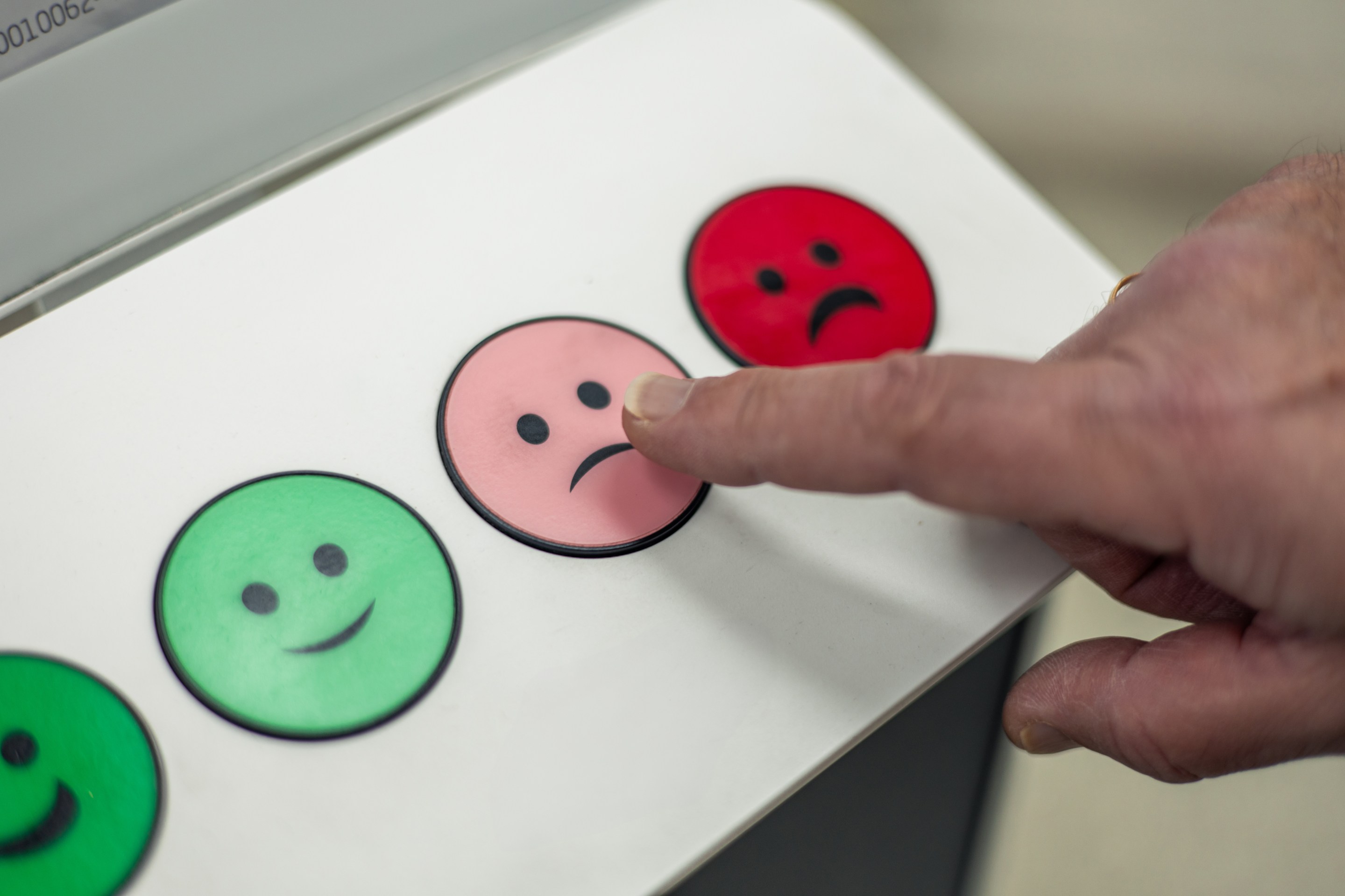 Hand pressing a slightly dissatisfied emoji button on a customer satisfaction feedback machine with green, pink and red smileys, expressing a neutral or somewhat negative experience closeup